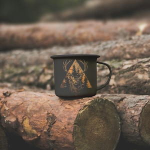 May include: A black enamel camping mug with a gold deer silhouette design inside a triangle. The mug is sitting on a stack of tree logs.