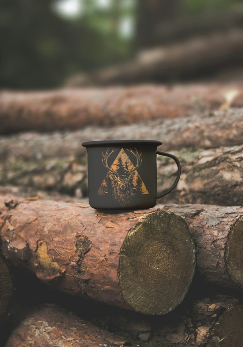 May include: A black enamel camping mug with a gold deer silhouette design inside a triangle. The mug is sitting on a stack of tree logs.