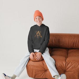 May include: A young person wearing a black hoodie with a graphic of a deer in a triangle, a light blue pair of pants, and a bright orange beanie. They are sitting on a brown leather couch with their legs crossed.