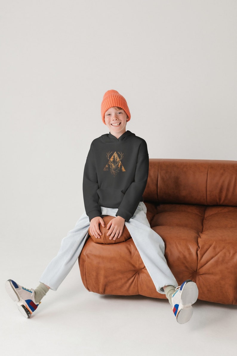 May include: A young person wearing a black hoodie with a graphic of a deer in a triangle, a light blue pair of pants, and a bright orange beanie. They are sitting on a brown leather couch with their legs crossed.