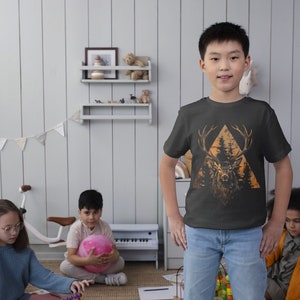 May include: A young boy wearing a black t-shirt with a graphic print of a deer in a forest scene. The graphic is in shades of orange, brown, and green. The boy is standing in a room with a white wood panel wall and a white piano in the background.