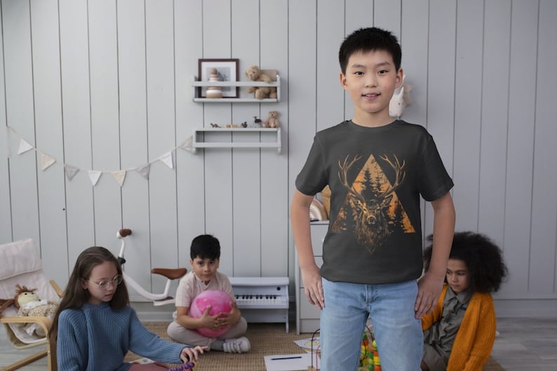 May include: A young boy wearing a black t-shirt with a graphic print of a deer in a forest scene. The graphic is in shades of orange, brown, and green. The boy is standing in a room with a white wood panel wall and a white piano in the background.