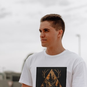 May include: A white t-shirt with a graphic print of a deer's head in a triangle shape with trees and a sunset behind it.