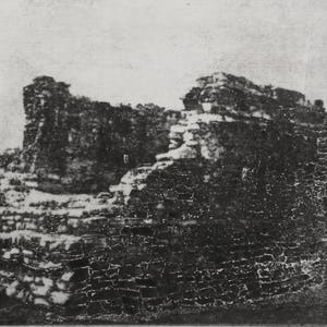 Puede incluir: Una impresión fotográfica en blanco y negro que representa las ruinas de piedra desgastadas de una estructura antigua. La imagen muestra muros de mampostería en capas con texturas visibles, bordes desmoronados y sombras oscuras.