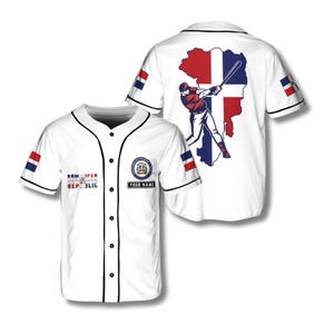 Pode incluir: Camisola de beisebol branca com acabamento preto, com as cores e imagens da República Dominicana. A frente exibe texto e um patch para personalização. A parte de trás mostra um jogador de beisebol rebatendo uma bola sobre um mapa da República Dominicana.