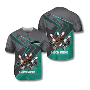 Puede incluir: Camiseta de béisbol gris con temática de bolos. El diseño presenta un bolo con sombrero de vaquero y sosteniendo dos pistolas. El texto "I'M ON STRIKE" está impreso en la parte delantera y trasera. La camiseta tiene una parte delantera abotonada y un diseño turquesa y gris.