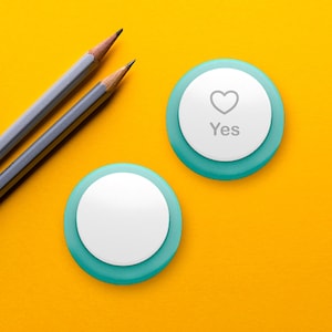 May include: Two gray pencils and two teal and white circular objects on a yellow background. One object has a heart symbol and the word "Yes" printed on it. The other object is blank.