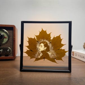 May include: A framed artwork featuring a silhouette of a woman's profile within a golden leaf design. The artwork is encased in a black frame with a clear backing, and is set against a soft, illuminated background. The piece is displayed on a wooden surface.