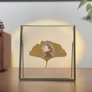 Peut inclure: Un cadre photo carré en métal doré avec un portrait de femme sur une feuille de ginkgo dorée. Le profil de la femme est en noir et blanc, et la feuille est placée sur un fond crème.
