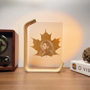 Op de afbeelding: Decoratieve lamp met een gedroogd esdoornblad met een vrouwenportret. Het blad is ingekapseld in een helder paneel, ondersteund door een gebogen houten frame. De lamp geeft een warm licht.