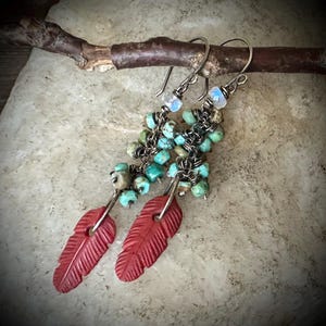 May include: Dangle earrings featuring dark metal wire chains adorned with small, round, variegated turquoise beads and a single iridescent moonstone bead at the top. Each earring terminates in a carved, matte red feather-shaped pendant.