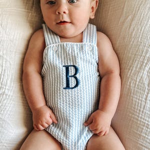 Könnte beinhalten: Ein Baby in einem blau-weiß gestreiften Strampler mit dem Buchstaben "B" auf der Vorderseite. Der Strampler hat Knopfriemen und ist aus Seersucker-Stoff. Das Baby liegt auf einem weißen Laken.