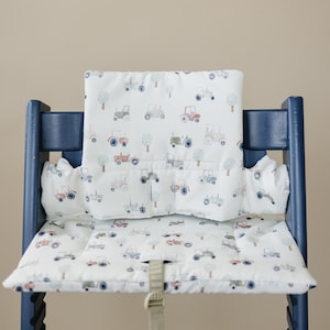 Puede incluir: Un juego de cojines blancos para trona con un estampado de tractores en azul, rojo y gris. El juego incluye un cojín para el asiento y una funda para el respaldo. La trona es azul oscuro.