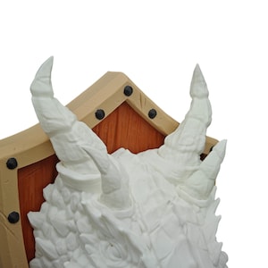 Puede incluir: Escultura de cabeza de drag&oacute;n blanca montada en un escudo de madera con marco beige. El escudo tiene remaches de metal negro alrededor de los bordes.