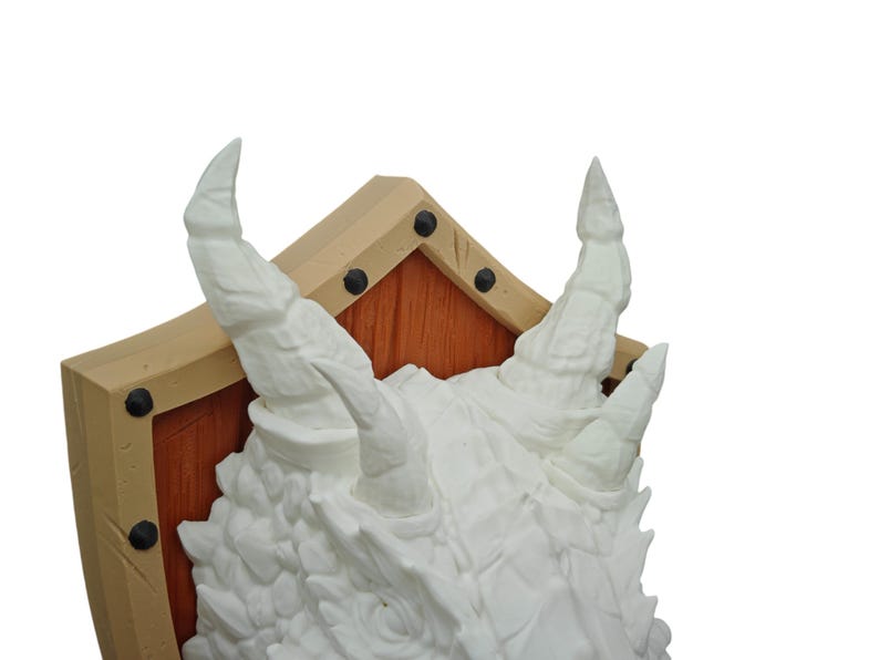 Puede incluir: Escultura de cabeza de drag&oacute;n blanca montada en un escudo de madera con marco beige. El escudo tiene remaches de metal negro alrededor de los bordes.