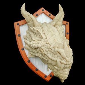 Puede incluir: Una escultura de cabeza de drag&oacute;n blanca montada en un escudo con borde naranja y remaches negros. El drag&oacute;n tiene una cara texturizada detallada y dientes afilados.