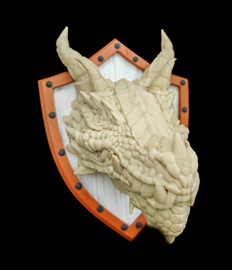 Puede incluir: Una escultura de cabeza de drag&oacute;n blanca montada en un escudo con borde naranja y remaches negros. El drag&oacute;n tiene una cara texturizada detallada y dientes afilados.