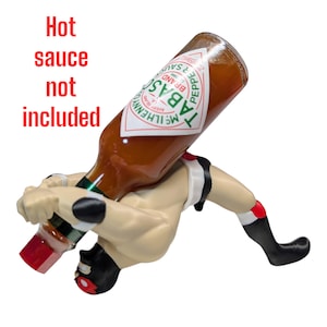 May include: A wrestler figurine holding a bottle of Tabasco hot sauce. The wrestler is in a wrestling pose, with one leg extended and the other bent. The bottle is upside down, with the cap facing the wrestler's head. The wrestler is wearing a white and red wrestling outfit. The bottle is labeled "TABASCO" in green letters.