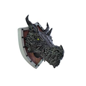 Puede incluir: Cabeza de drag&oacute;n negra montada en un escudo de madera con detalles plateados. El drag&oacute;n tiene una piel texturizada y escamosa y una expresi&oacute;n amenazante.