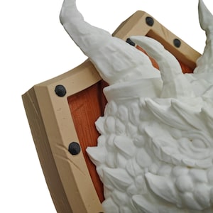 Puede incluir: Una escultura de cabeza de drag&oacute;n blanca montada en un escudo de madera. La cabeza del drag&oacute;n est&aacute; detallada con escamas y un cuerno afilado. El escudo est&aacute; pintado de marr&oacute;n con detalles negros.