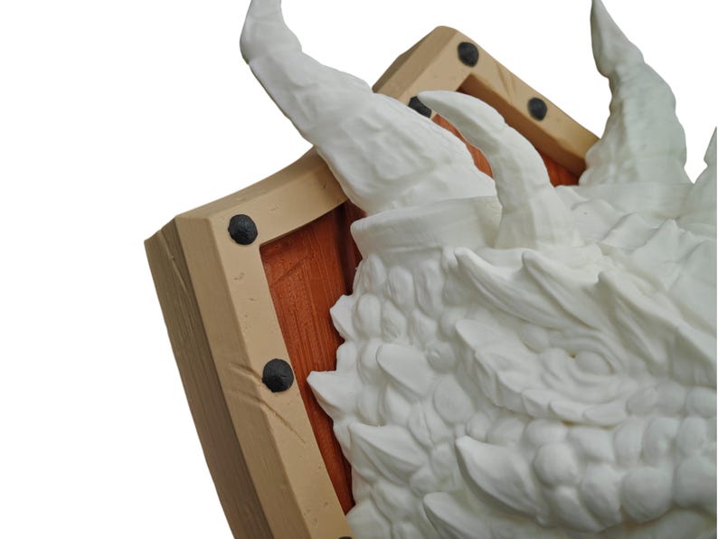 Puede incluir: Una escultura de cabeza de drag&oacute;n blanca montada en un escudo de madera. La cabeza del drag&oacute;n est&aacute; detallada con escamas y un cuerno afilado. El escudo est&aacute; pintado de marr&oacute;n con detalles negros.