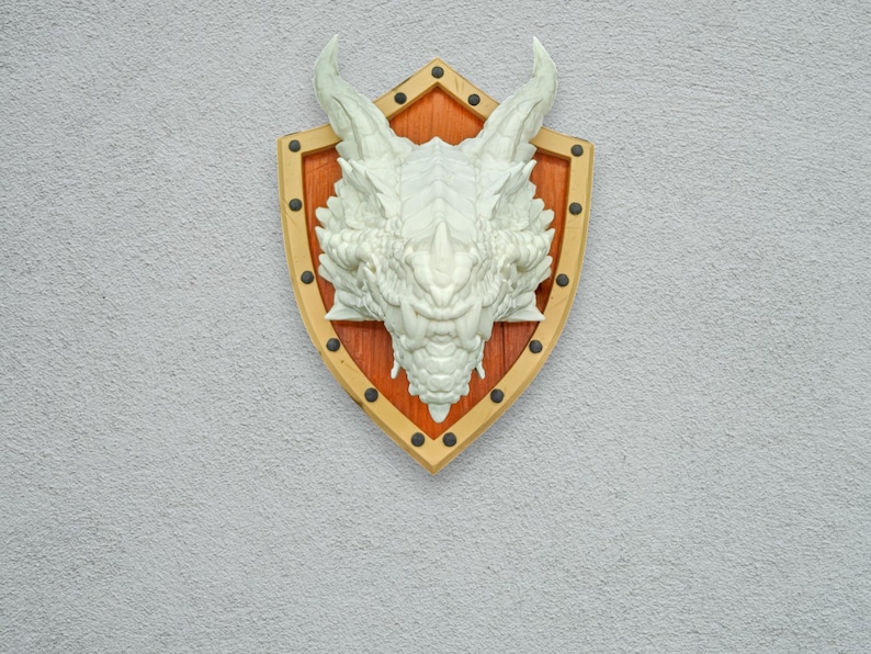 Puede incluir: Una escultura de cabeza de drag&oacute;n blanca montada en un escudo de madera con fondo marr&oacute;n y detalles dorados.