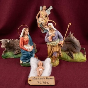 Pode incluir: Um conjunto de presépio com figuras de uma mulher ajoelhada em azul e vermelho, um homem de pé em roxo e bege com um cajado, um bebé em tecido branco, um anjo, uma vaca e um burro. Uma etiqueta de madeira exibe o texto Nacimiento Hebreo 74.75€.