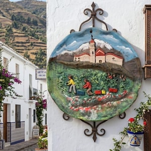 Puede incluir: Una placa de pared circular de cerámica pintada a mano que presenta una escena en relieve de figuras cosechando en un campo verde bajo una iglesia blanca con techo rojo y una montaña nevada, montada sobre un marco decorativo de hierro forjado.