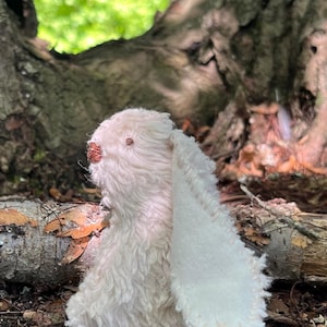 May include: A small, off-white plush rabbit toy with a shaggy, textured fabric body and a long, smooth ear. The rabbit features a small, dark brown stitched nose and a single visible eye, crafted with visible seam detailing along the ear edge.