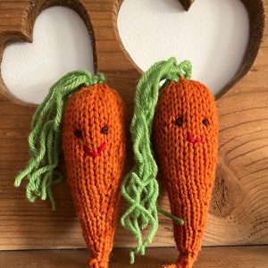 May include: Two knitted orange carrots with green yarn tops and smiling faces. The carrots are on a wooden background with heart-shaped cutouts.