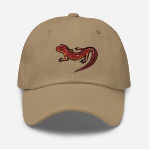 Può includere: Un cappello da baseball color beige con un salamandra rosso e arancione ricamato sul davanti.