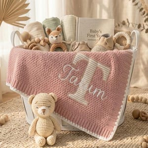Könnte beinhalten: Ein weißer Korb gefüllt mit Babyartikeln, darunter eine rosa Strickdecke mit dem Namen Tatum und einem großen gestickten Buchstaben T, ein beiger Häkel-Teddybär, eine Fuchsrassel, Mulltücher, ein Erinnerungsbuch und kleine Babyschuhe.