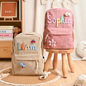 Peut inclure: Deux sacs à dos en velours côtelé, un beige et un rose, sont présentés. Le sac à dos beige porte le nom "Liam" en lettres colorées et des appliqués de voiture et d'avion. Le sac à dos rose présente le nom "Sophia" et un appliqué de fleur. Les deux ont des poches avant.
