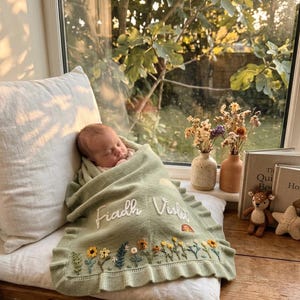 Könnte beinhalten: Eine salbeigrüne Strickdecke für Babys mit gerüschtem Rand, bestickt mit dem Namen Fiadh Violet in weißer Schreibschrift und verziert mit einem bunten Blumenmuster, das Sonnenblumen und einen kleinen Regenbogen enthält.