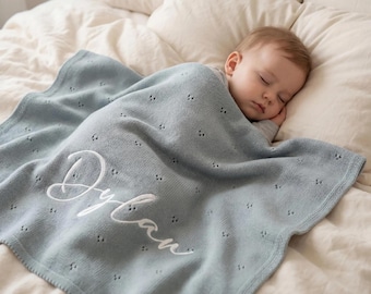May include: A light blue knitted baby blanket featuring a delicate pointelle pattern and the name Dylan embroidered in white cursive script. The blanket is draped over a sleeping infant who is resting on a soft, cream-colored bedding surface.
