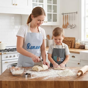 May include: A light blue and white pinstriped apron with the name Renee embroidered in blue, and a black and white pinstriped apron with the name Miko embroidered in multi-coloured letters, both featuring adjustable neck straps and front pockets.