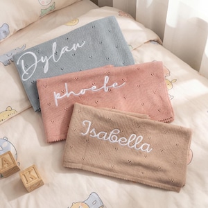 Puede incluir: Tres mantas rectangulares de punto en azul, rosa empolvado y beige, cada una con un delicado patrón de calados y nombres bordados en blanco: Dylan, phoebe e Isabella. Las mantas están superpuestas sobre una superficie estampada.