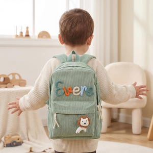 May include: A light green corduroy backpack with the name "Evere" embroidered in colorful letters. The backpack features a small front pocket and a lion applique. The backpack is designed for children.