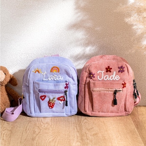 May include: Two small corduroy backpacks with embroidered names. The purple backpack says "Lara" and the pink backpack says "Jade".