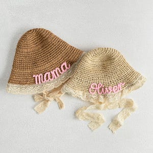 May include: Two straw sun hats. One is brown with the word "mama" in pink script. The other is beige with the name "Oliver" in pink script. Both hats have a lace trim and ribbon ties.