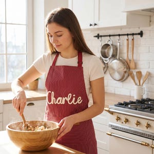 May include: A dark red apron with thin white vertical stripes and the name Charlie embroidered in white script across the chest. The apron features a neck strap and a front pocket, worn over a plain white short-sleeved t-shirt.