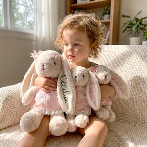 Könnte beinhalten: Drei cremefarbene Plüschhasen mit langen Schlappohren, die jeweils ein rosa Kleid mit weißer Spitzenborte tragen. Die Ohren sind mit den Namen Cecilia, Ava und Laiya in Schreibschrift bestickt. Jeder Hase hat ein kleines rosa Schleifenaccessoire.