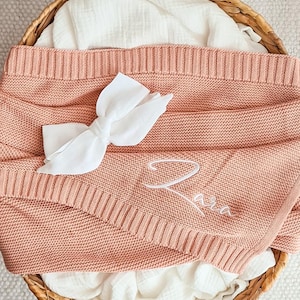 Può includere: Una copertina per neonati in maglia rosa cipria con un fiocco bianco e il nome "Aria" ricamato in bianco.