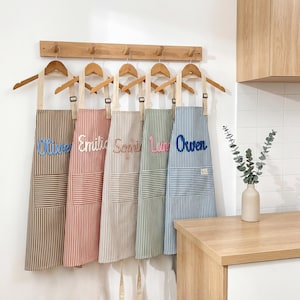 Könnte beinhalten: Fünf gestreifte Schürzen in Braun, Rot, Beige, Grün und Blau hängen an einer Holzstange. Jede Schürze hat einen anderen Namen: Oliver, Emilia, Sophie, Lun und Owen. Die Schürzen haben Taschen und hängen in einer Küche.