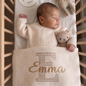 Könnte beinhalten: Ein schlafendes Kleinkind in einem cremefarbenen Rippenpullover, das in einem Holzbettchen mit einem kleinen gestrickten Teddybären liegt. Eine cremefarbene Decke bedeckt das Kind, verziert mit einem großen beigen Vichy-Karo-E und dem Namen Emma.