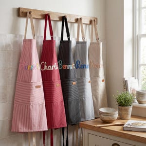 Könnte beinhalten: Fünf gestreifte Schürzen in verschiedenen Farben, darunter Rosa, Rot, Grau und Beige, hängen an einer Holzstange. Jede Schürze ist mit einem Namen bestickt. Die Schürzen haben eine Tasche und einen Lederaufnäher.