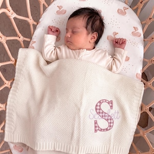 Könnte beinhalten: Eine cremefarbene Strickdecke für Babys mit einem großen, gestickten Buchstaben S in einem rosa-weißen Vichy-Muster, wobei der Name Scarlett in Schreibschrift unter dem Buchstaben eingestickt ist, während sie auf einem schlafenden Säugling liegt.