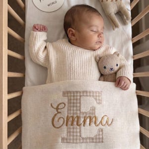 Könnte beinhalten: Ein schlafender Säugling in einem cremefarbenen Rippenpullover, der unter einer cremefarbenen Strickdecke liegt, die mit einem großen beigen Vichy-Karo-Buchstaben E und dem in goldener Schreibschrift gestickten Namen Emma verziert ist.