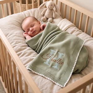 Könnte beinhalten: Eine salbeigrüne Strickdecke für Babys mit weißem Bogenrand, verziert mit einem großen hellblau-weiß karierten Buchstaben H und dem in brauner Schreibschrift gestickten Namen Hallie, eingewickelt um ein schlafendes Baby neben einem Stofftier.