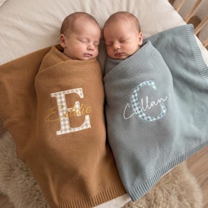 Könnte beinhalten: Zwei Säuglinge in gestrickten Decken. Eine Decke ist hellbraun mit einem großen, weiß-hellbraun karierten Buchstaben E und dem in Gold gestickten Namen Eddie. Die andere Decke ist salbeigrün mit einem blau-weiß karierten Buchstaben C und dem in Weiß gestickten Namen Callan.